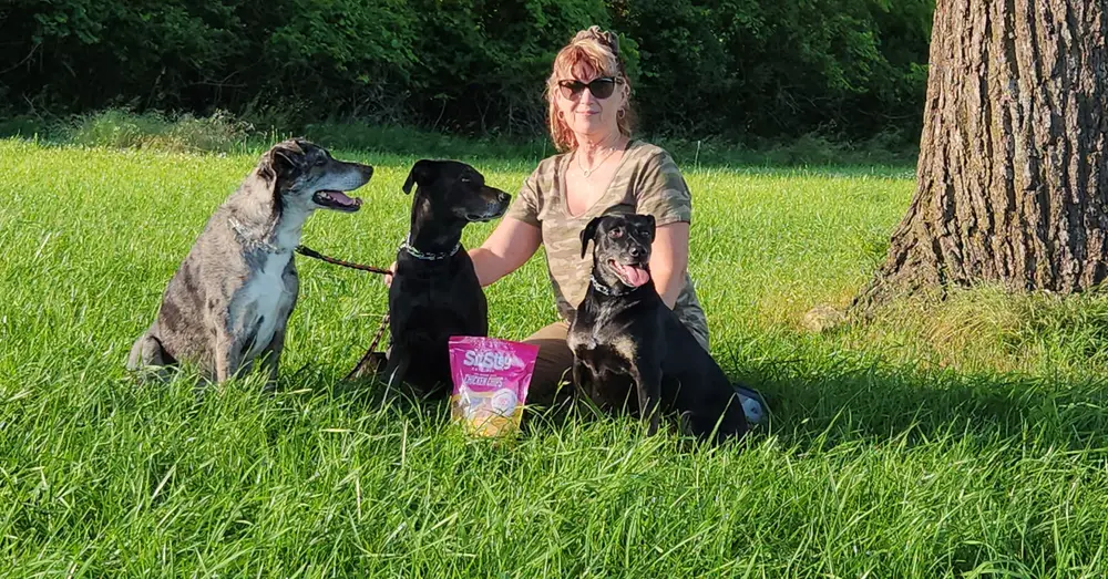 Jodi with her three dogs and a bag of SitStay Chicken Chips in a sunny field