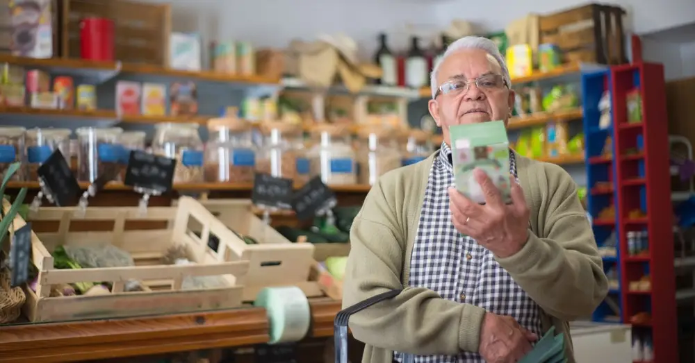 Elderly man examines product in grocery store aisle, representing daily shopping routine.