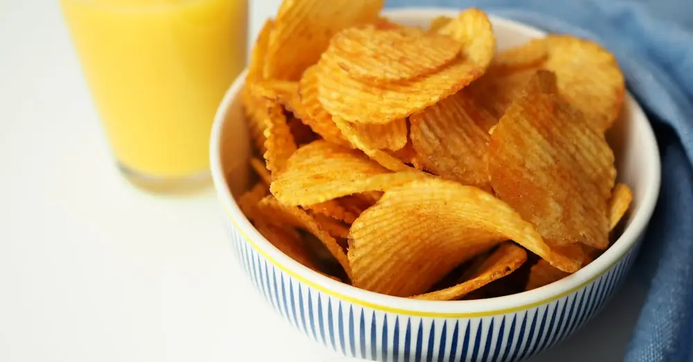 Close-up of crispy potato chips in a bowl with fresh orange juice on a white surface.