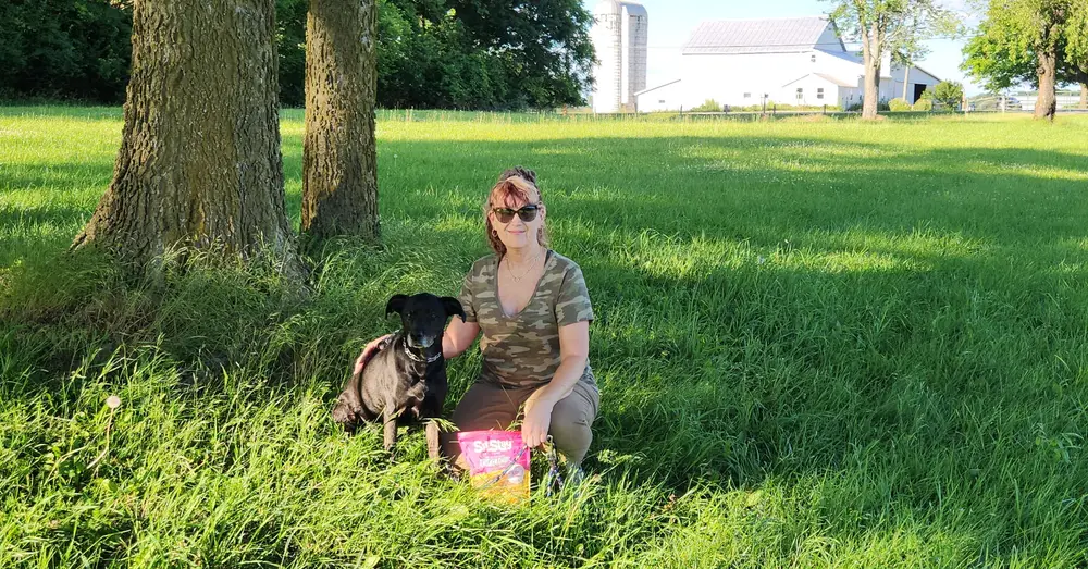 Jodi and a dog in a farm field with a bag of SitStay Chicken Chips