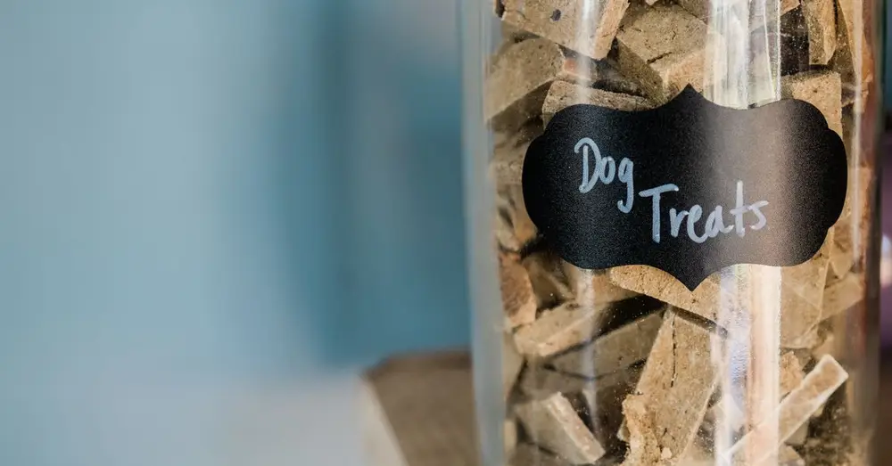 A close-up image of dry dog treats stored in a labeled glass jar on a wooden surface.