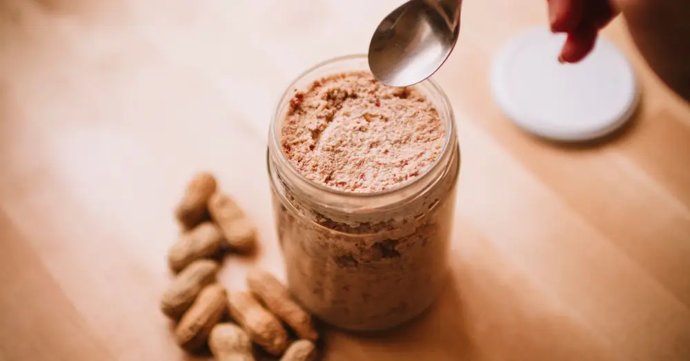 A glass jar of homemade peanut butter on a wooden table with peanuts and a spoon.