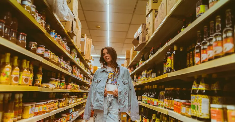 Woman in denim browsing grocery store aisle with products on shelves.